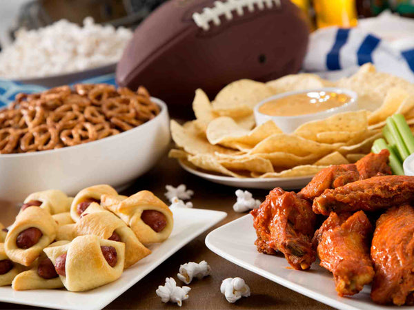 A tailgate spread of wings, brats, sliders and dip laid out on a table beside a grill
