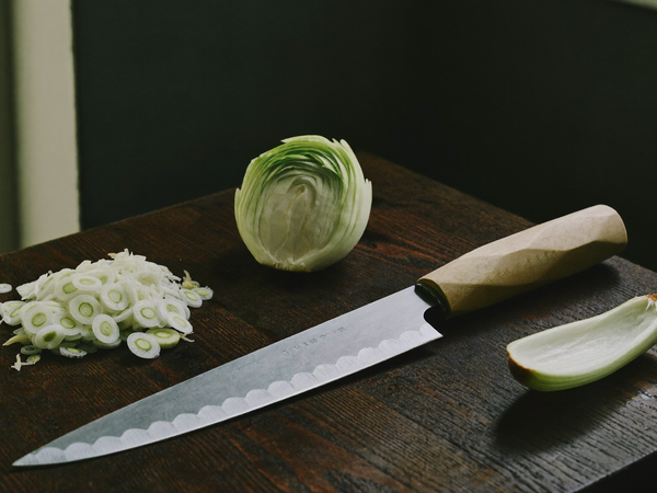 Hand-forged Japanese chef knife with octagonal maple handle on a walnut cutting board beside sliced scallions and daikon