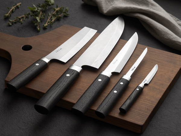 Japanese kitchen knife set laid out in a row on a dark walnut cutting board in soft window light